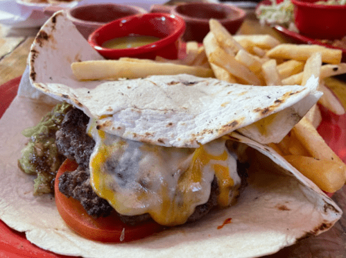 A burger with melted cheese and tomato wrapped in a tortilla, served with fries and dipping sauces on the side.